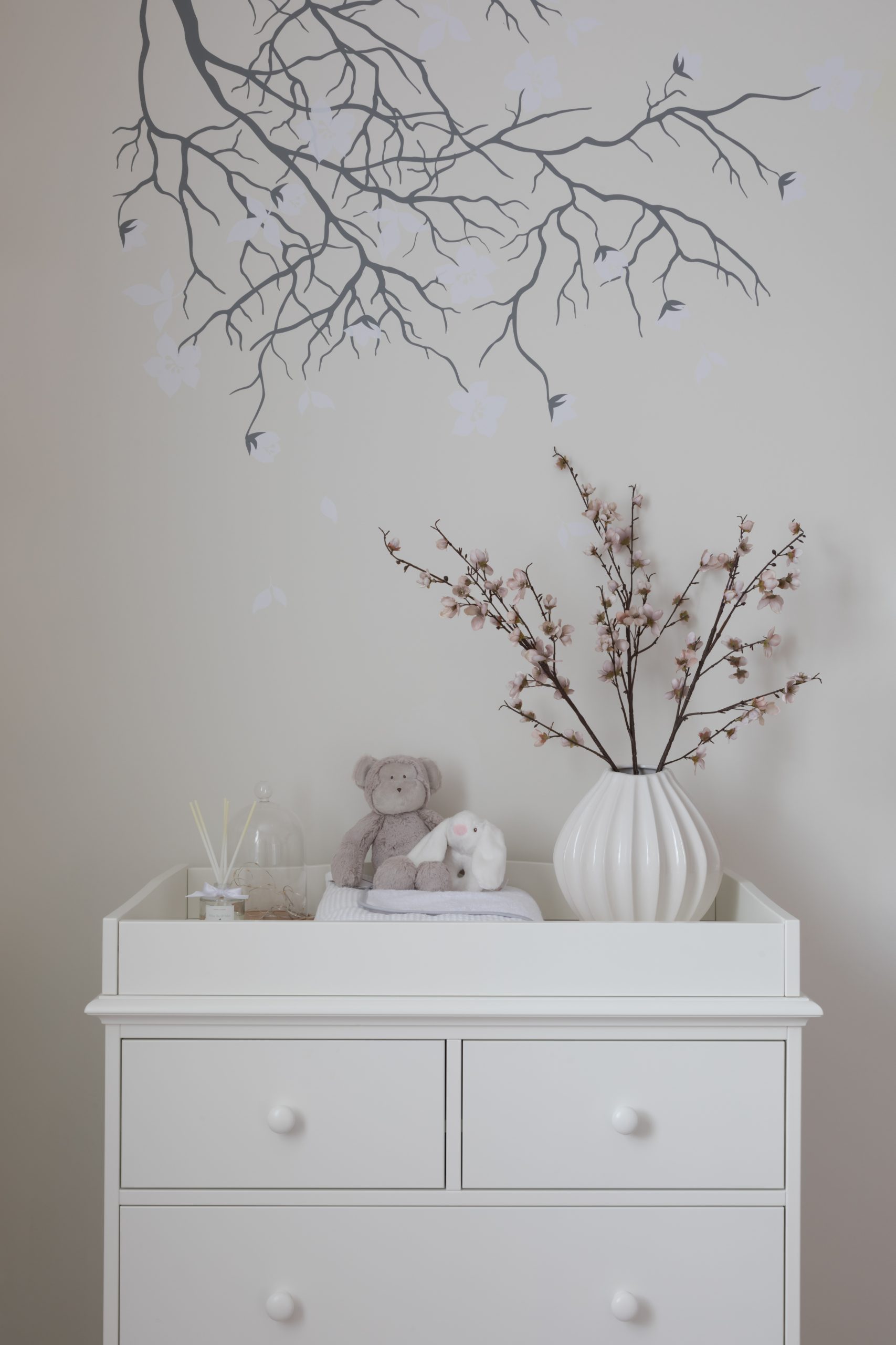 Side chest of draws with children's toys on top with flower pot and small pink flowers in side. Wall art of tree branches with white flowers behind.