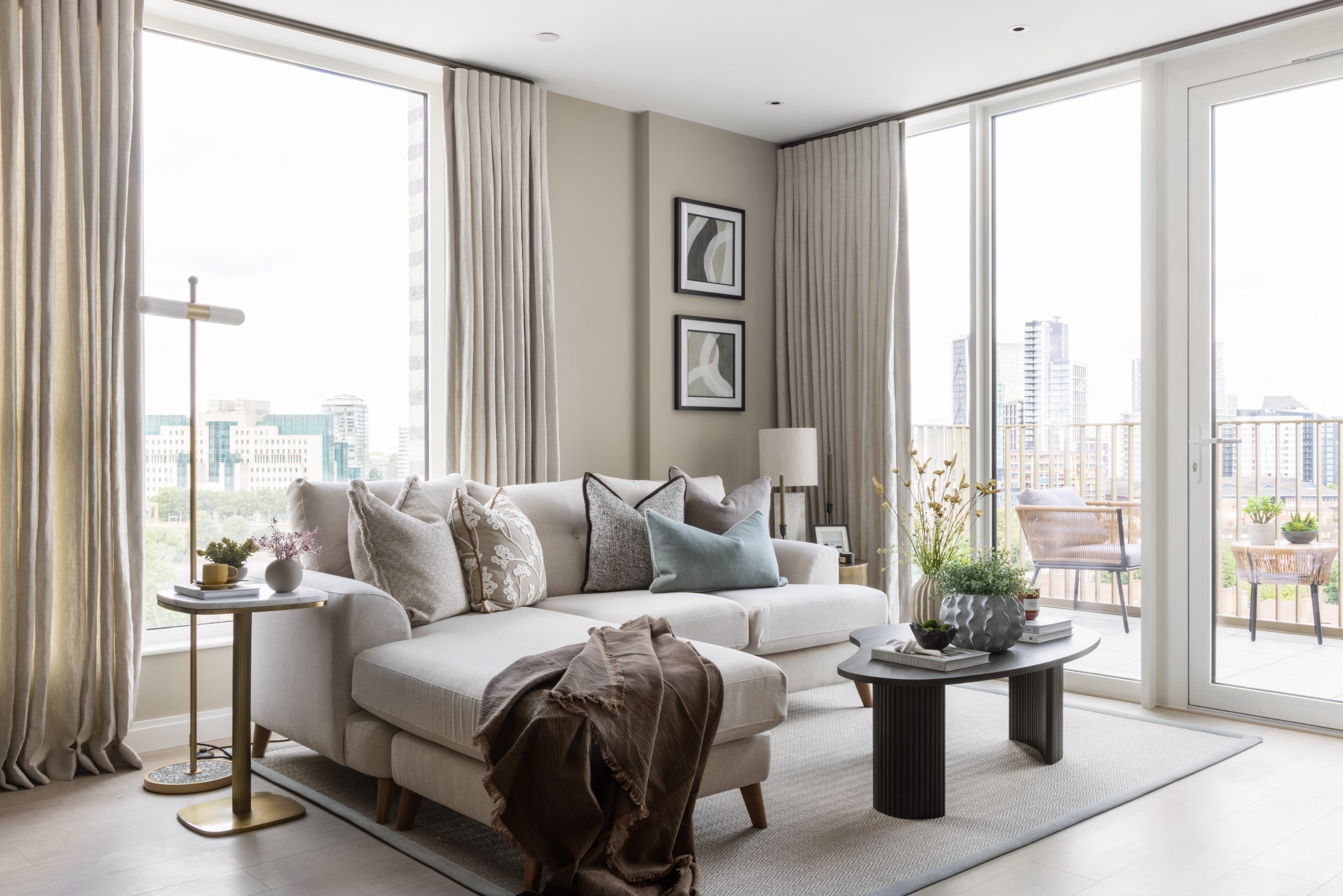 Oval Village Apartment living room space with cream corner sofa and view of the balcony with cushioned seating and table and various large buildings in background.