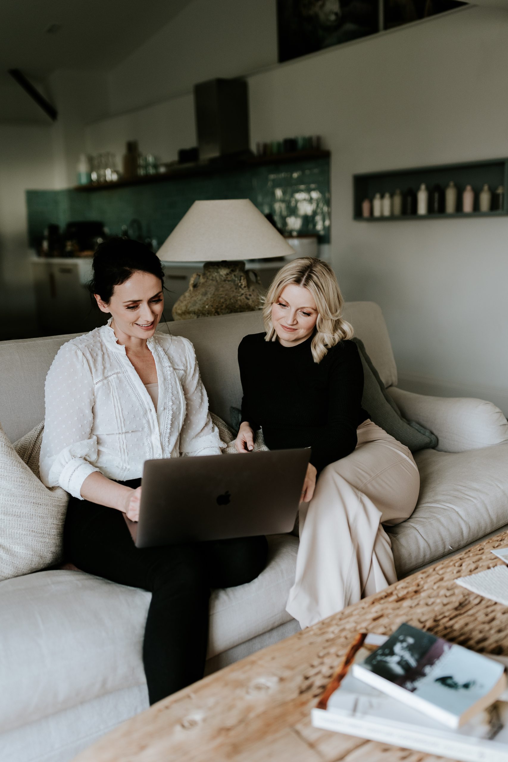 Emily and Danielle (high end interior designers) sitting side by side on a cream sofa with laptops and fabric swatches to hand