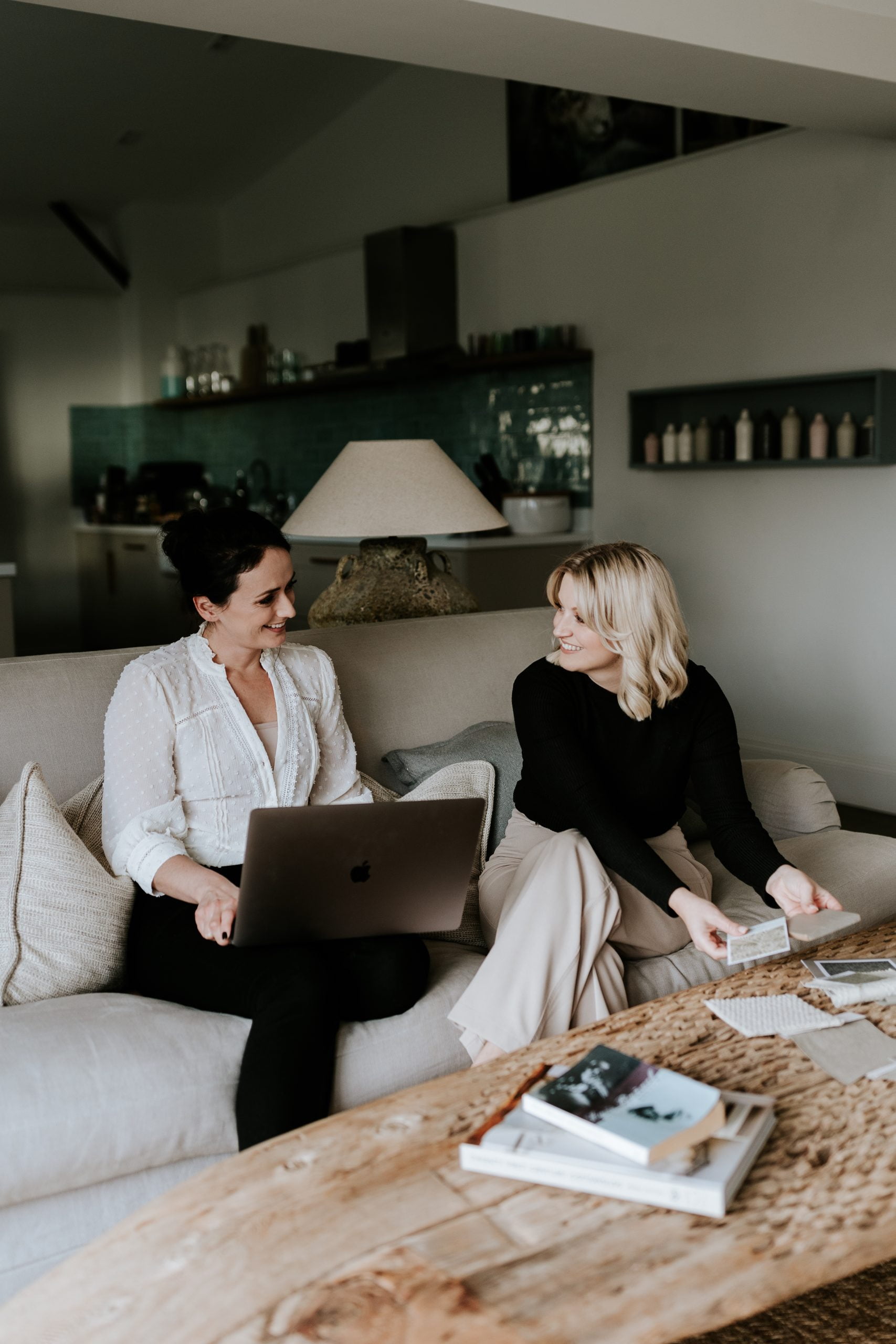 Emily and Danielle (high end interior designers) sitting side by side on a cream sofa with laptops and fabric swatches to hand