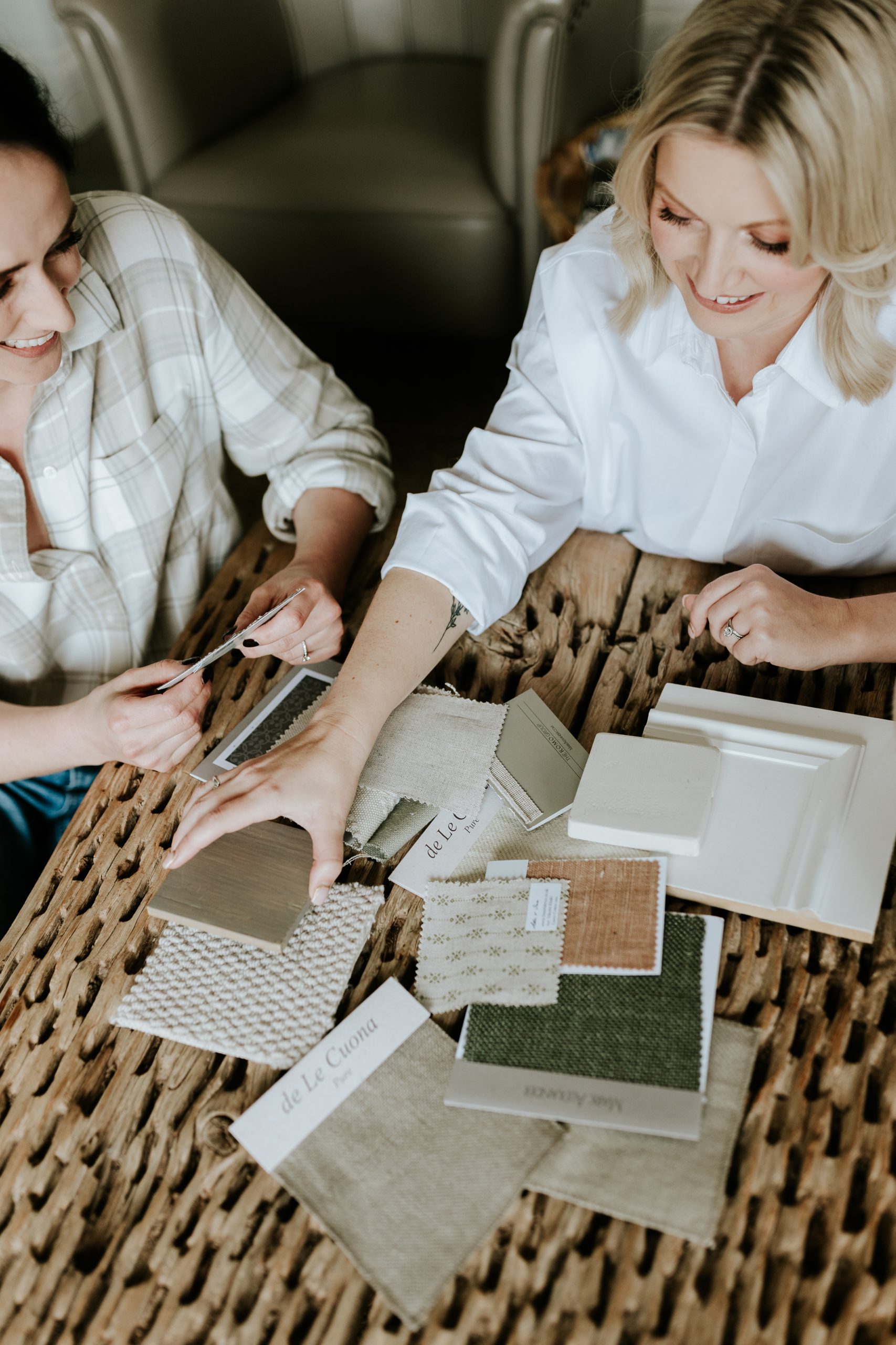 Emily in white shirt reaching for swatch on textured effect table while sitting next to danielle in light coloured flannel shirt | High end interior design studio